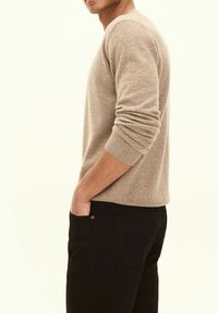 Hellbrauner Strickpullover mit langen Ärmeln und gerippten Bündchen, kombiniert mit dunkelgrauten Hosen, die ein sichtbares Taschendetail und einen subtilen Akzent aufweisen.