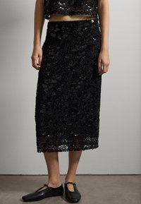 Zwarte midi-rok van kant met een verhoogd bloemenpatroon, versierd met pailletten. Past goed bij een zwart cropped top en zwarte platte schoenen.