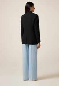 Blazer nero su misura con una sola fessura sul retro, abbinato a pantaloni ampi di colore azzurro. Tessuto liscio e design strutturato.