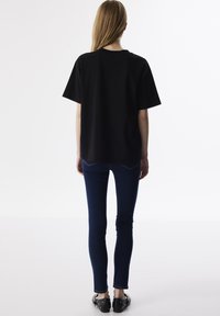 Zwarte oversized T-shirt met korte mouwen, weergegeven vanuit de achterkant. Gecombineerd met blauwe skinny jeans en zwarte slip-on schoenen met studs.