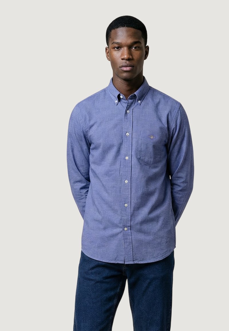 Jeune homme debout, les mains derrière le dos, portant une chemise bleue à manches longues boutonnée et un pantalon bleu foncé sur un fond uni.