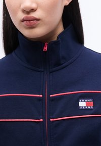 Κοντινό πλάνο ατόμου που φοράει μπλε ναυτικό μπουφάν με φερμουάρ και ροζ διακοσμητική ραφή, με το λογότυπο Tommy Jeans στο αριστερό στήθος.