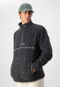 Fuzzy gray fleece pullover with a half-zip collar, green patterned band across the chest, and side pockets. Complemented by a green cap.