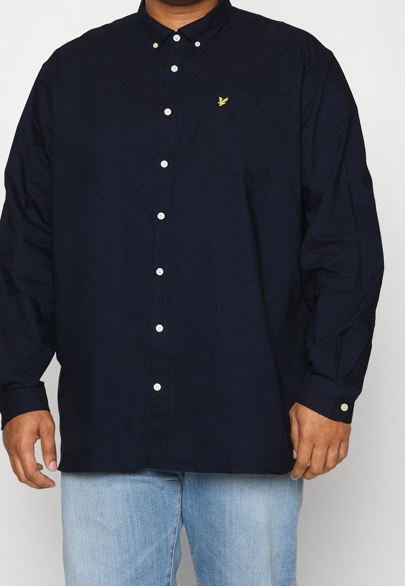 Homme portant une chemise bleu marine foncé à manches longues avec des boutons blancs et un jean bleu clair, debout devant un fond uni.