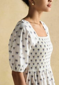 Rochie albă cu model floral albastru, decolteu pătrat, corset plisat și mâneci voluminoase. Materialul pare ușor și texturat.