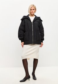 Zwarte pufferjack met een grijze fleece-voering capuchon, ritsluiting en vo pockets; gedragen over een lichtgekleurde, knie-lange jurk.