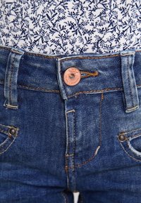 Blue denim jeans feature a classic design with a copper button and visible stitching. The fabric shows slight fading and has metal accents.