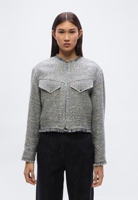 Σύντομο γκρι σακάκι από τουίντ με φερμουάρ εμπρός, δύο τσεπάκια στο στήθος, ξεφτισμένες άκρες και στρογγυλή λαιμόκοψη. Φορεμένο με μαύρο υφασμάτινο παντελόνι.