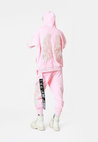 Sudadera con capucha rosa y joggers a juego con bordado de alas, que presentan una etiqueta prominente en los pantalones y botas blancas gruesas.