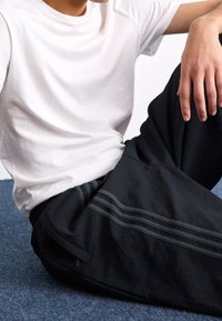 Svarta texturerade byxor med tre mörkgrå ränder längs sidorna, kombinerade med en enfärgad vit t-shirt, modell sittande på en blå yta.