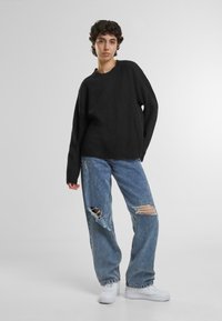 Schwarzer, übergroßer Pullover mit geripptem Saum, kombiniert mit hellblauen, verwaschenen Jeans mit großen Rissen an den Knien und weißen Sneakers.