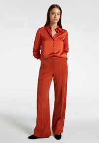 Camicia in raso a maniche lunghe con bottoni di un vivace arancione, abbinata a pantaloni a gamba larga in corda a coste dello stesso colore e scarpe nere.