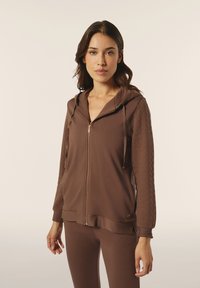 Felpa con zip marrone e maniche testurizzate, caratterizzata da una vestibilità rilassata, cappuccio regolabile con cordino e un discreto dettaglio del logo. Abbinata a pantaloni coordinati.