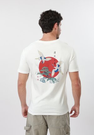 Mężczyzna widziany od tyłu, ubrany w białą koszulkę z nadrukiem dwóch żurawi, drzewa oraz czerwonego słońca z pionowymi azjatyckimi znakami.