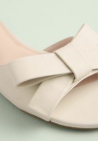 Lys beige åben tå sko med en tekstureret læderfinish; har en stor sløjfedetalje over forfoden og en blød lyserød indersål.
