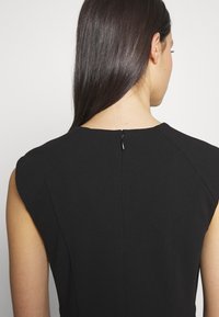 Femme aux longs cheveux foncés portant une robe noire sans manches avec une fermeture éclair dans le dos, sur un fond clair uni.
