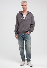 Grå zip-hoodie med teksten "BIG TAG AUTHENTIC", parret med lyseblå jeans og hvide sneakers, som har en afslappet pasform og snøre i hætten.