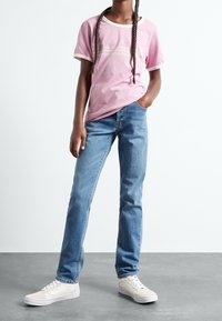 Niña con trenzas largas, vistiendo una camiseta rosa, jeans azules y zapatillas blancas, de pie sobre un suelo gris contra un fondo blanco.