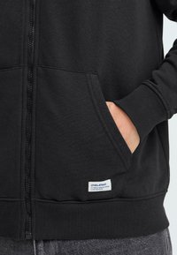 Sweat à capuche zippé noir en tissu doux, avec poches latérales, poignets côtelés et une petite étiquette de marque près de l'ourlet.