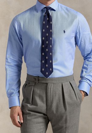 Homme portant une chemise bleu clair, une cravate bleu marine à motif et un pantalon gris plissé taille haute, les mains dans les poches.