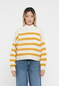 Crèmekleurige trui met gele strepen, een losse pasvorm, geribde mouwen en onderrand. Gecombineerd met hoog-geritste blauwe jeans.