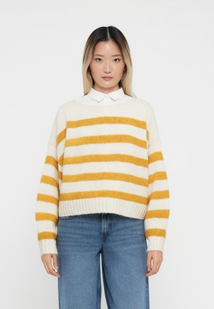 Crèmekleurige trui met gele strepen, een losse pasvorm, geribde mouwen en onderrand. Gecombineerd met hoog-geritste blauwe jeans.