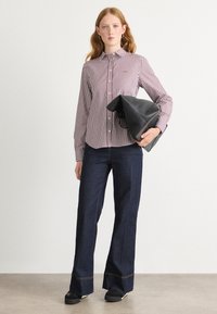 Camicia a righe rossa e bianca, abbinata a jeans a gamba larga blu scuro. Il modello tiene una borsa nera, caratterizzata da un design elegante.