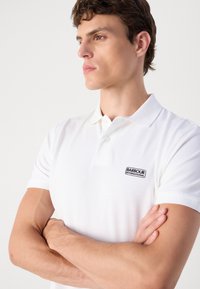 Fehér pólóing pamutból, rövid ujjakkal, a bal mellrészen fekete Barbour International logóval. Sima textúra.