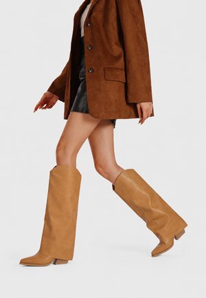 Donna che cammina indossando stivali in pelle color cuoio fino al ginocchio, minigonna nera, giacca marrone con bottoni e top bianco su uno sfondo uniforme.