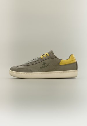 Scarpa da ginnastica bassa grigia con toppa gialla sul tallone e lacci, suola color crema e logo "REPLAY" sul lato, mostrata su sfondo neutro.