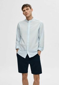 Camicia azzurra chiara a maniche lunghe con colletto alla coreana, abbinata a pantaloni corti scuri. La camicia presenta maniche arrotolate e una vestibilità comoda.
