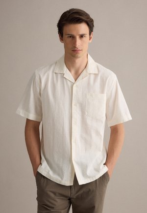 Joven con cabello corto y oscuro, lleva una camisa blanca de manga corta abotonada y pantalones marrones, de pie con las manos en los bolsillos.