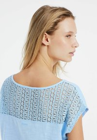 Lichtblauw topje met een achterkant van kantpatroon, ronde halslijn en korte mouwen. Gladde katoenen voorkant contrasteert met de gestructureerde kanten achterkant.