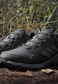 Chaussures de randonnée noires Adidas avec semelles texturées sur le sol de la forêt avec de l’herbe et des feuilles en arrière-plan.