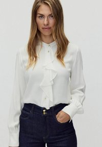 Vit blus med rynkad framsida och långa ärmar, kompletterad med gyllene knappar. I kombination med mörka jeans.