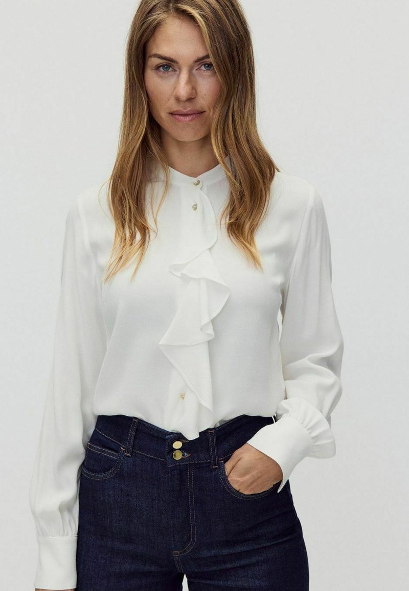 Vit blus med rynkad framsida och långa ärmar, kompletterad med gyllene knappar. I kombination med mörka jeans.