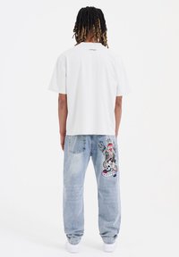 Camiseta blanca de manga corta con un logo bordado en la parte trasera, combinada con unos jeans de mezclilla azul claro que presentan un colorido bordado en la pierna.