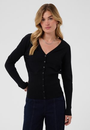 Femme aux cheveux blonds ondulés portant un cardigan noir boutonné, un jean foncé et un collier en argent, debout devant un fond clair uni.
