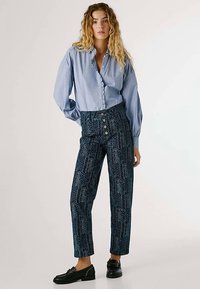 Svetlo modra srajca s črtami in dolgimi rokavi, kombinirana s temno modrimi vzorčastimi hlačami z visokim pasom, ki imajo vrstico gumbov. Črne loafer čevlje.