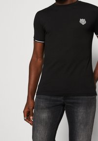 Camiseta de algodón negra con mangas cortas, que presenta un logo de tigre blanco en el pecho y ribetes blancos en las mangas. Combinada con unos jeans oscuros de mezclilla.