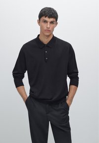 Schwarzer Pullover mit Polo-Kragen, langen Ärmeln und einer Knopfleiste mit drei Knöpfen. Geschmeidige Textur, lockere Passform, kombiniert mit dunklen Hosen.