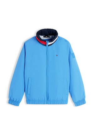 Dreng smilende iført en blå jakke, rød polo, beige shorts, hvide sokker og beige sneakers, stående med hænderne i lommerne.
