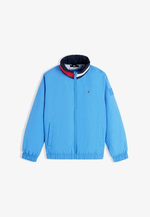 Jongen glimlachend met een blauwe jas, rood poloshirt, beige korte broek, witte sokken en beige sneakers, staand met handen in de zakken.