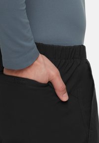 Zwarte shorts met elastische tailleband en een zijzak, gedragen met een nauwsluitend, grijs T-shirt met lange mouwen. De hand rust in de zak.