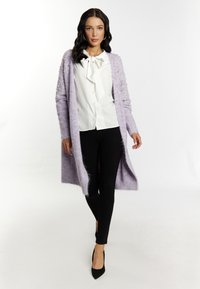 Cárdigan de punto lavanda con acentos de perlas en las mangas, llevado sobre una camisa blanca y pantalones negros, con diseño de abertura frontal y mangas largas.