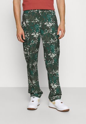 Uomo che indossa pantaloni cargo mimetici verdi e bianchi, scarpe da ginnastica bianche con suole marroni e una maglietta rossa a maniche corte.