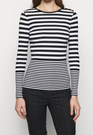 Femme portant une chemise à manches longues rayée horizontale noire et blanche avec des boutons dorés sur une épaule et un pantalon noir à fines rayures.