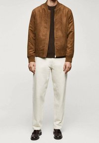 Blouson aviateur en daim marron, fermeture éclair, poignets côtelés, pull en maille marron en dessous, pantalon de couleur crème et chaussures en cuir noir.