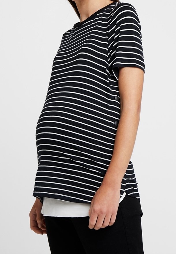 Personne enceinte portant une chemise à manches courtes noire et blanche rayée et un pantalon noir, se tenant devant un fond uni.