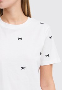 White cotton t-shirt with short sleeves, featuring small black embroidered bows evenly spaced across the fabric.